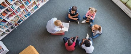 Students gathered around a person reading a book in a library setting.