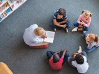 Students gathered around a person reading a book in a library setting.